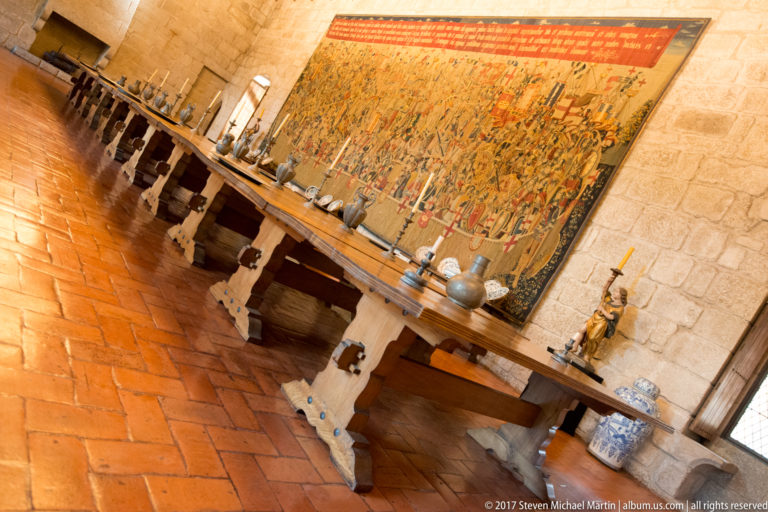 Angled view along a heavy wooden table set with plates, jugs, and candlesticks beneath a large narrative tapestry.