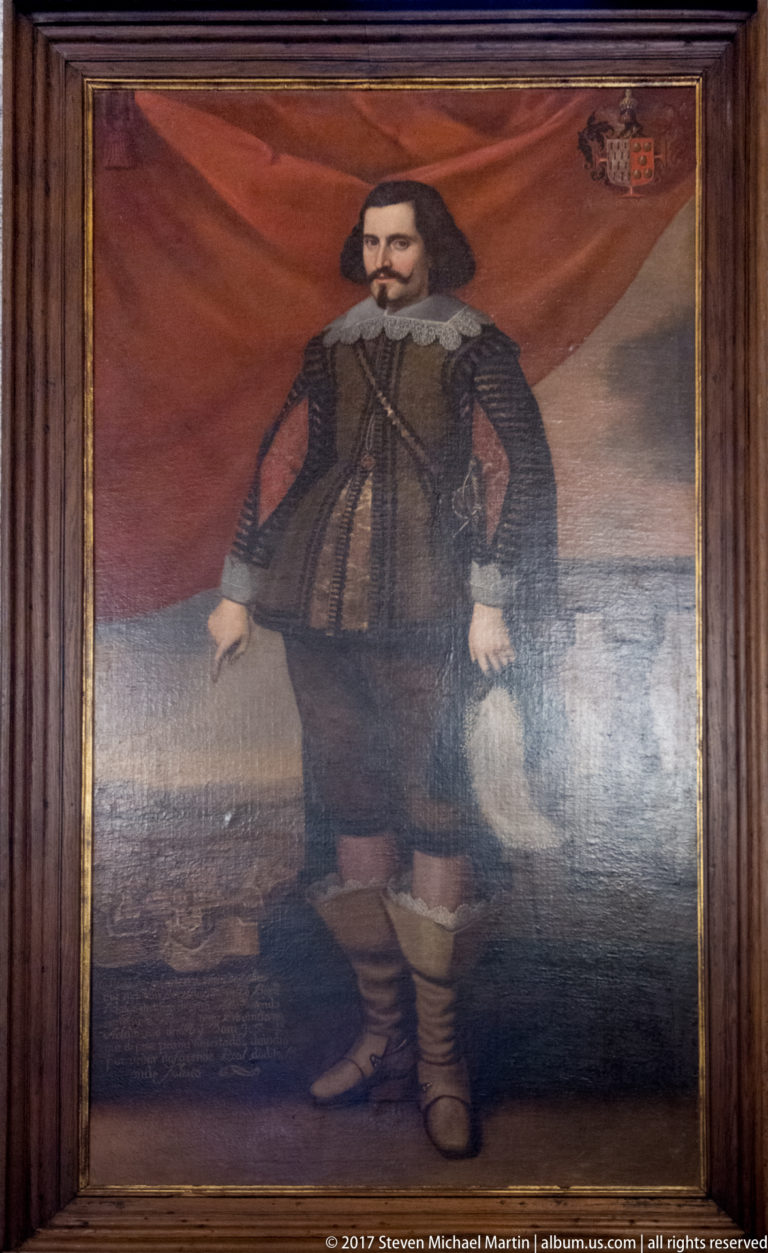 Full-length portrait of a dark-haired nobleman in black seventeenth-century dress and boots before red drapery, with a small coat of arms.