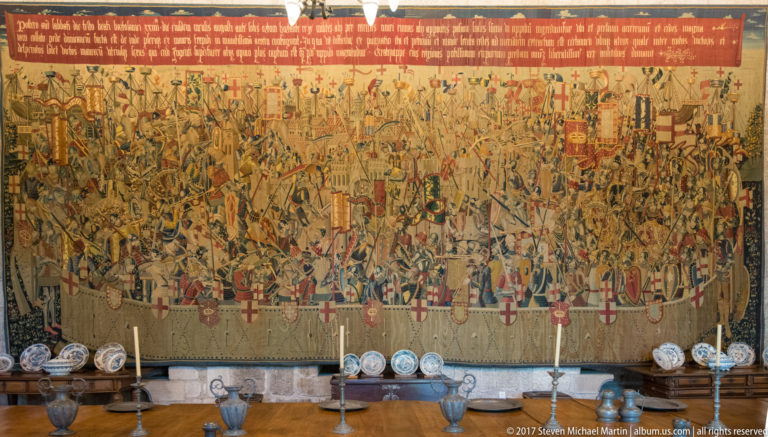 Large military narrative tapestry with soldiers, banners, and ships above rows of blue-and-white plates, jugs, and candlesticks.