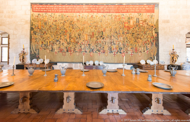 Wide wooden banquet table set with plates, vessels, and candlesticks before a monumental tapestry crowded with military figures.