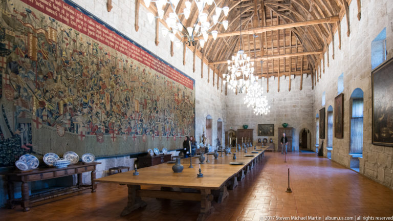 Long palace hall with exposed timber roof, chandeliers, central banquet table, tapestry and ceramics at left, and paintings at right.