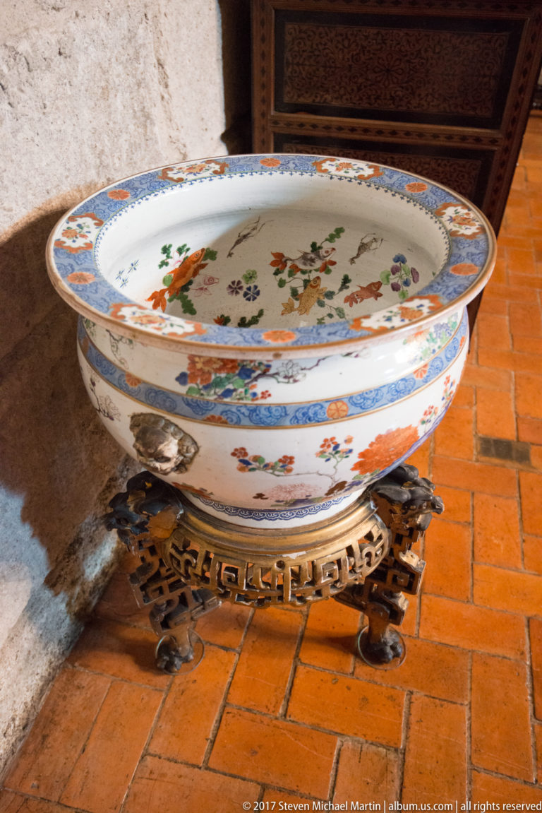 Large shallow porcelain basin painted with colorful birds and flowers, resting on an ornate dark metal stand over terracotta tiles.