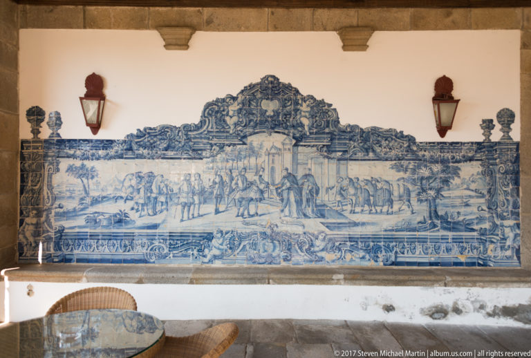 Large blue-and-white narrative azulejo mural with many figures and architecture, crowned by an ornate crest and flanked by lanterns.