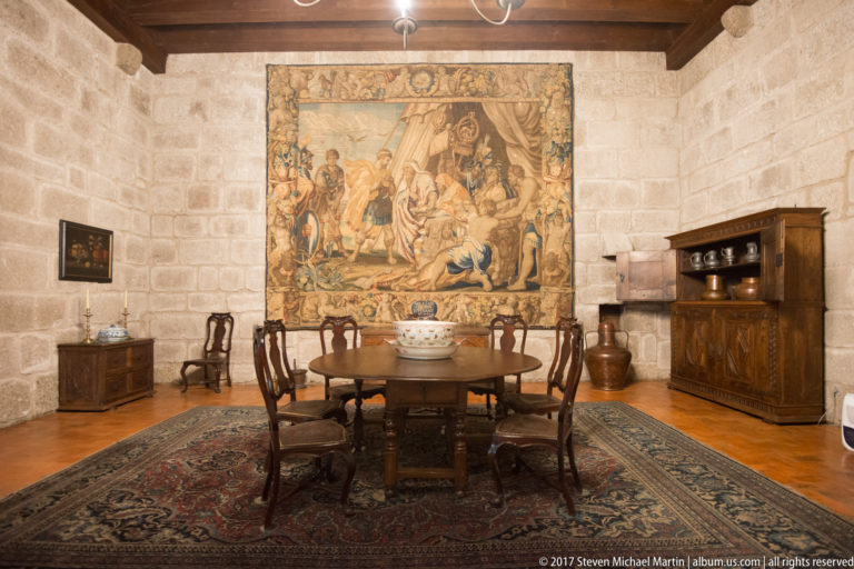 Round wooden dining table and carved chairs on a patterned carpet before a large figurative tapestry in a granite palace room.