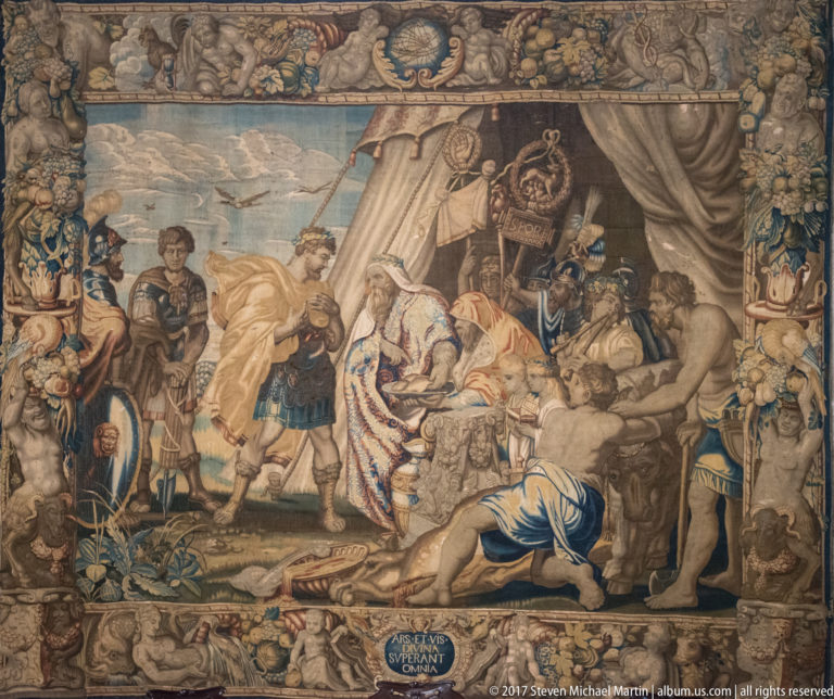 Large historic tapestry showing richly dressed figures, attendants, animals, and architectural details within an elaborate decorative border.