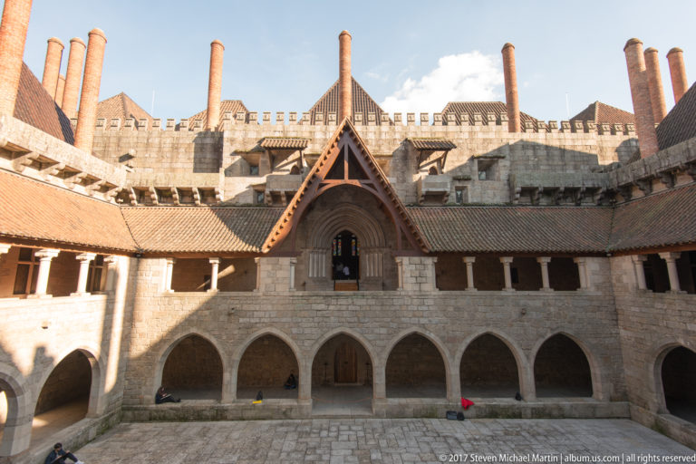 Granite palace courtyard with two-story arcades, steep roof, projecting gable, crenellations, and several tall cylindrical brick chimneys.