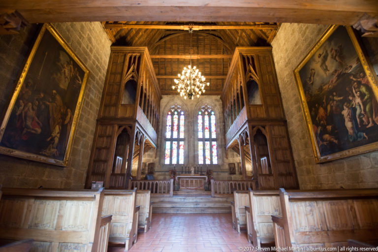 Palace chapel interior with wooden pews, side galleries, large religious paintings, chandelier, altar, and tall stained-glass windows.