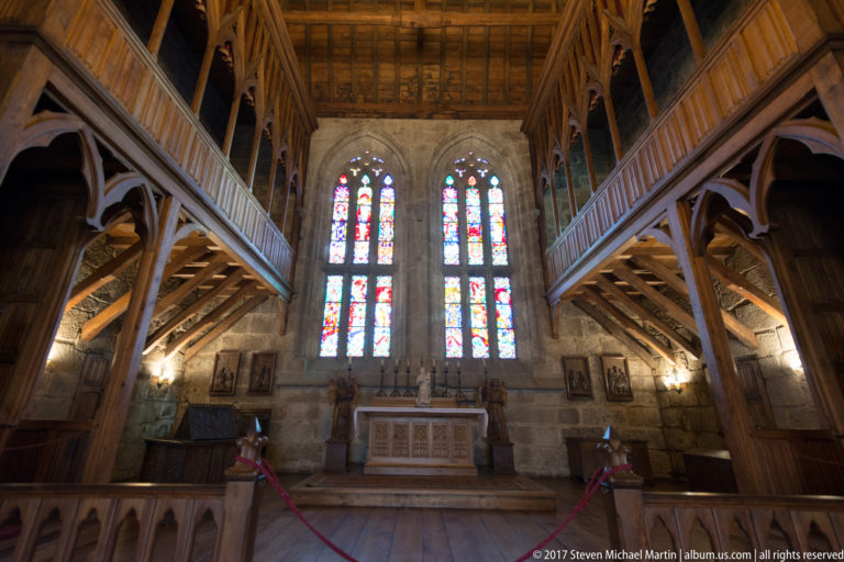 Altar beneath two tall stained-glass windows, framed by dark wooden galleries and heavy braces in a granite chapel interior.