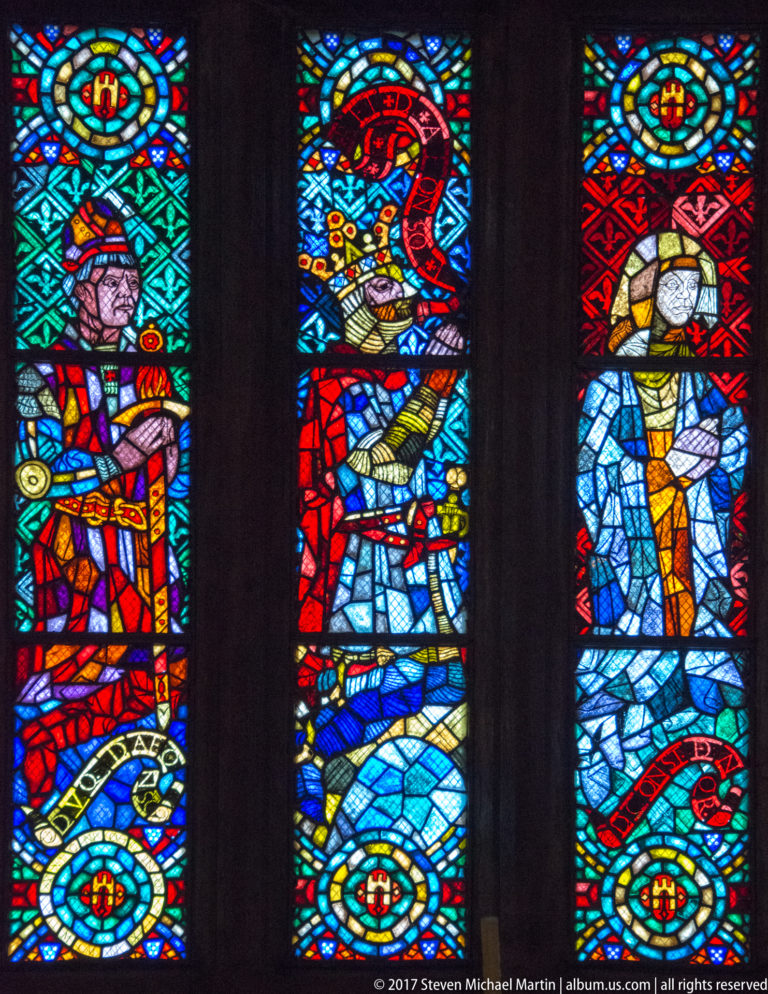 Three narrow stained-glass panels with stylized religious figures in bright red, blue, green, and gold geometric glass.