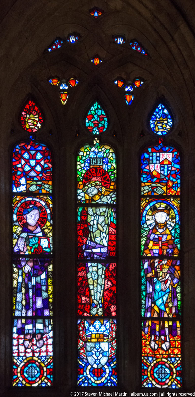 Three tall stained-glass lancets showing sacred figures in colorful robes beneath pointed arches, with small colored lights above.