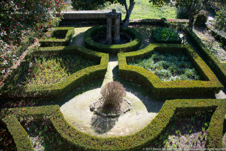 Formal garden viewed from above, with clipped boxwood parterres arranged around a small circular fountain and stone garden feature.