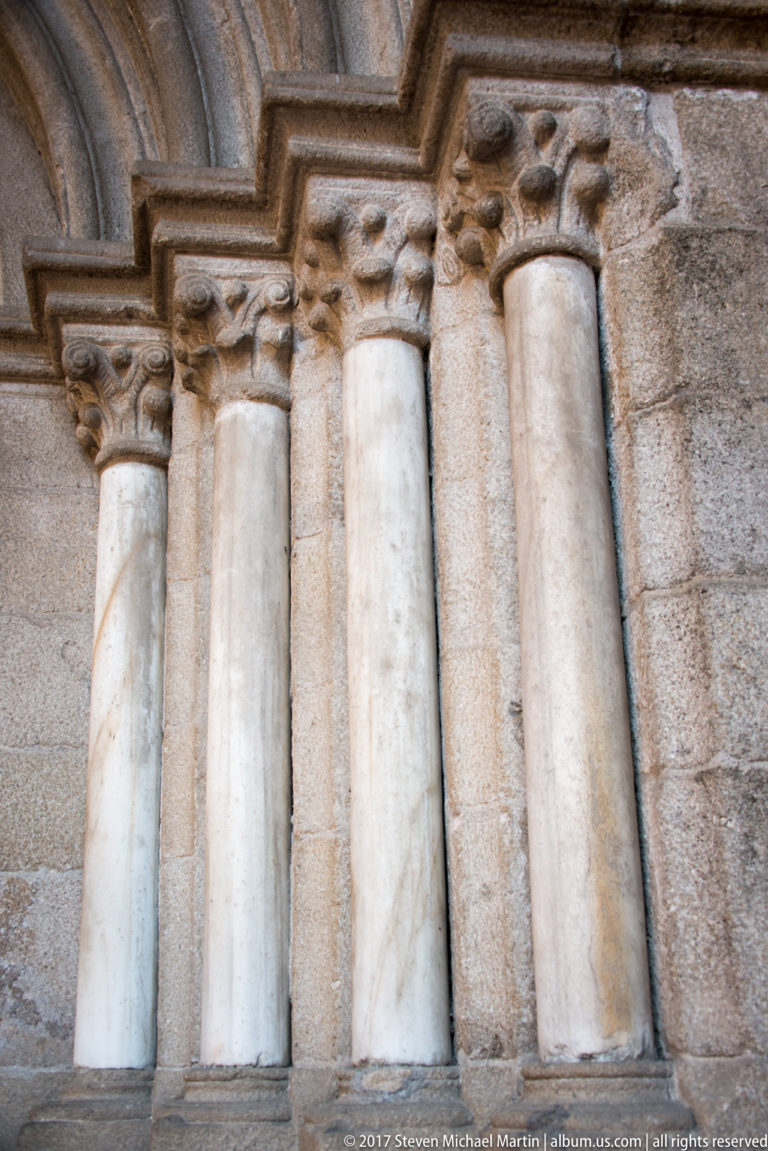 Close view of four pale stone colonnettes with carved capitals beneath stepped arch moldings in a rough granite wall.