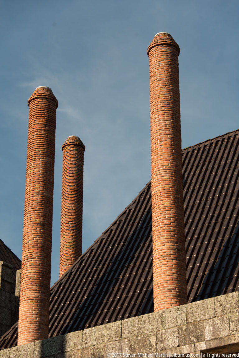 Four tall cylindrical brick chimneys rising above a steep tiled roof and granite wall against a blue sky.
