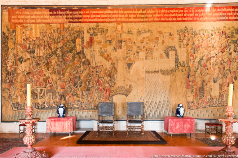 Monumental tapestry of a fortified city under attack, with cavalry and soldiers, behind two high-backed chairs and ceremonial furnishings.