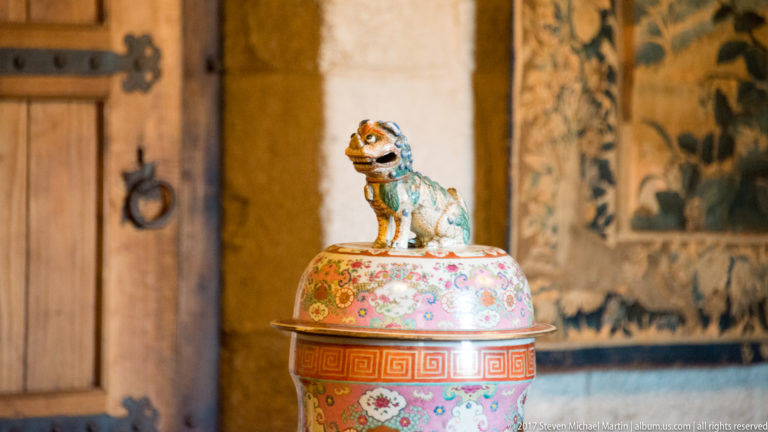 Small colorful guardian-lion figure standing atop the domed lid of a polychrome porcelain jar, with tapestry blurred behind.