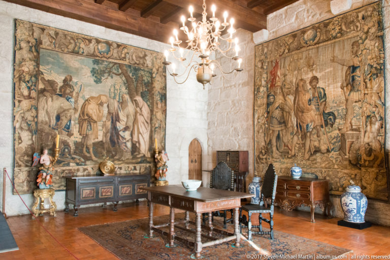 Historic granite chamber with two large tapestries, chandelier, carved table, high-backed chairs, cabinets, and porcelain vessels.