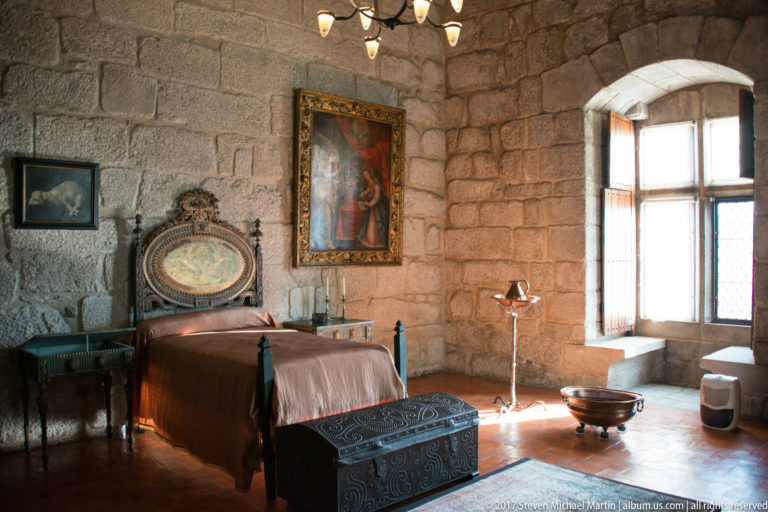 Historic stone bedchamber with wooden bed, ornate headboard, patterned chest, framed painting, side tables, and sunlit window.