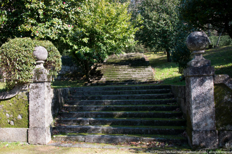 Weathered stone garden staircase rising between masonry posts with ball finials, clipped shrubs, trees, and moss-covered walls.