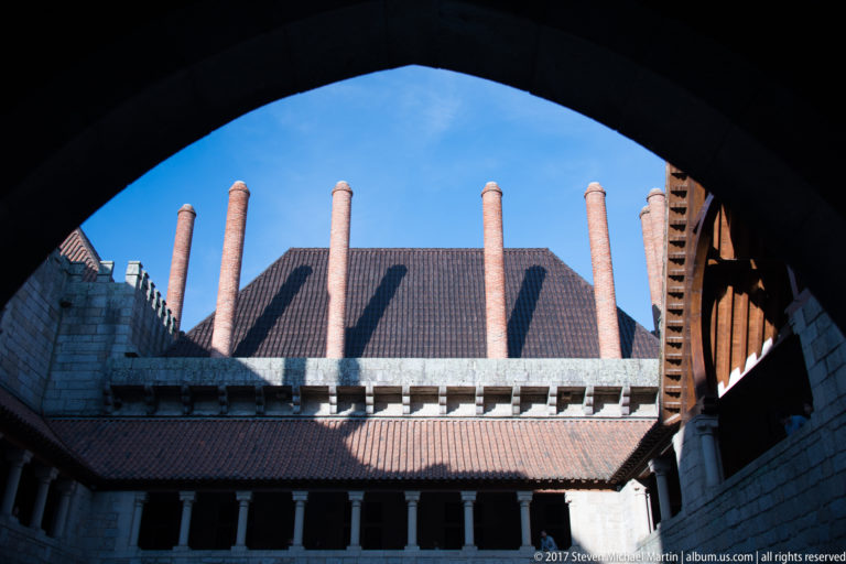 Palace courtyard and tall brick chimneys seen through a dark arch, with arcaded galleries, steep roofs, and blue sky beyond.