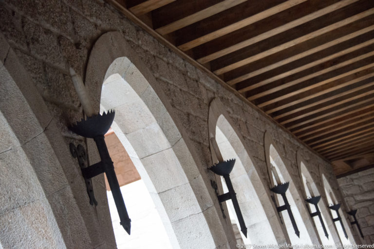 Receding granite arcade beneath a timber ceiling, with repeated pointed arches and black wrought-iron torch brackets on the piers.