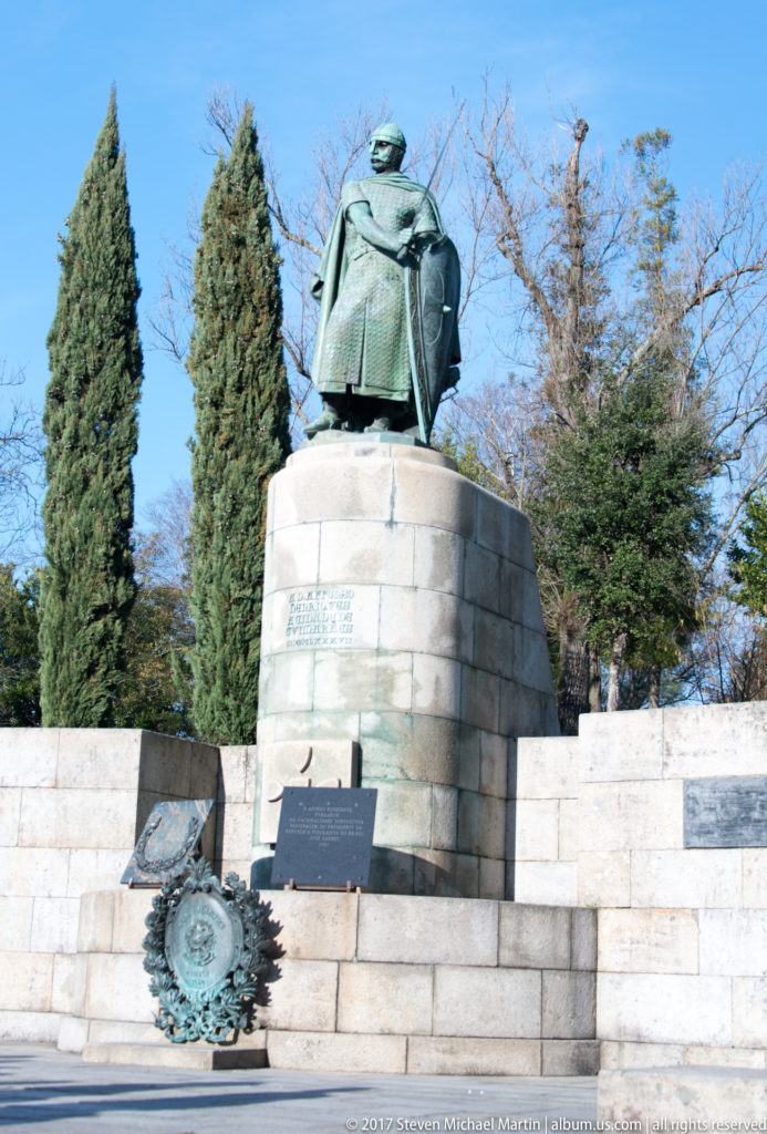Bronze statue of Afonso Henriques in armor with sword and shield atop a pale-stone pedestal, surrounded by memorial plaques and trees.