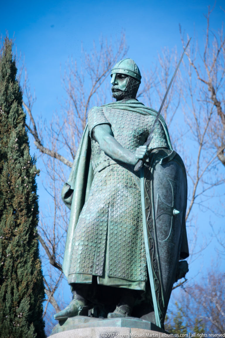 Close low-angle view of bronze Afonso Henriques statue wearing armor and helmet while holding a sword and large decorated shield.
