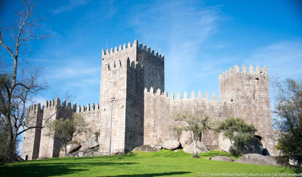 Guimarães Castle on a green slope, with tall crenellated keep, curtain walls, lower towers, granite masonry, and large foreground boulders.