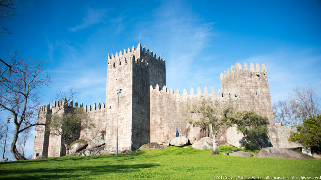 Wide view of Guimarães Castle across green lawn, with tall keep, crenellated towers, curtain walls, boulders, and leafless trees.