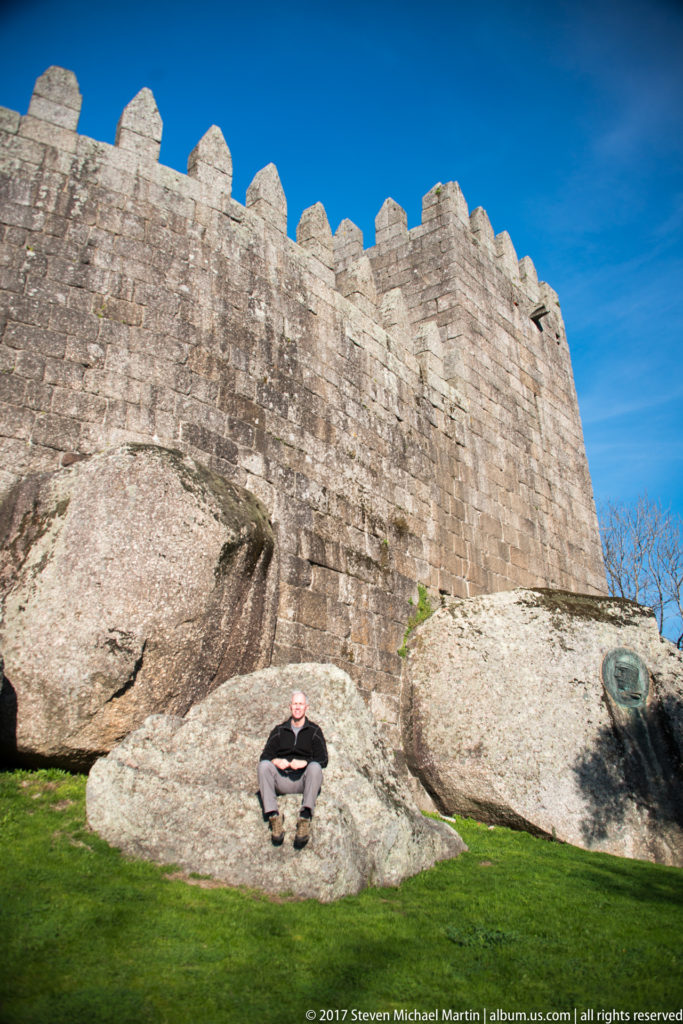 Man seated on a granite boulder below the high crenellated stone walls of Guimarães Castle under a vivid blue sky.