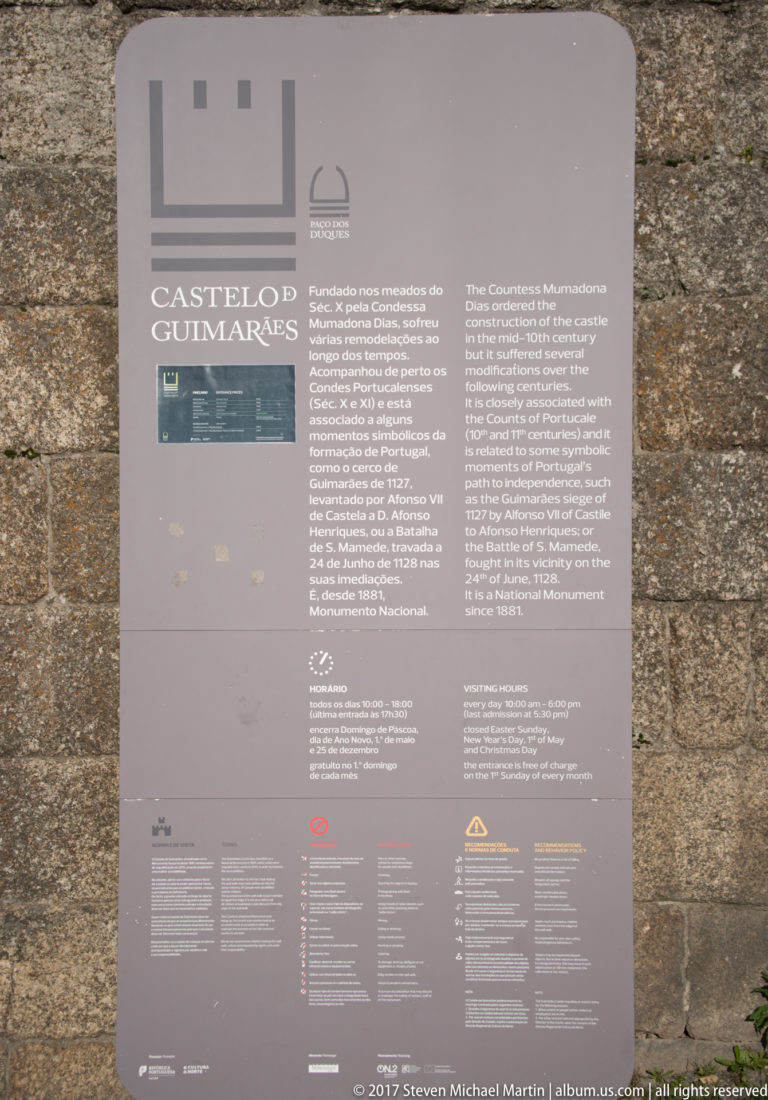 Gray bilingual visitor information panel headed “Castelo de Guimarães,” mounted against rough stone castle masonry.