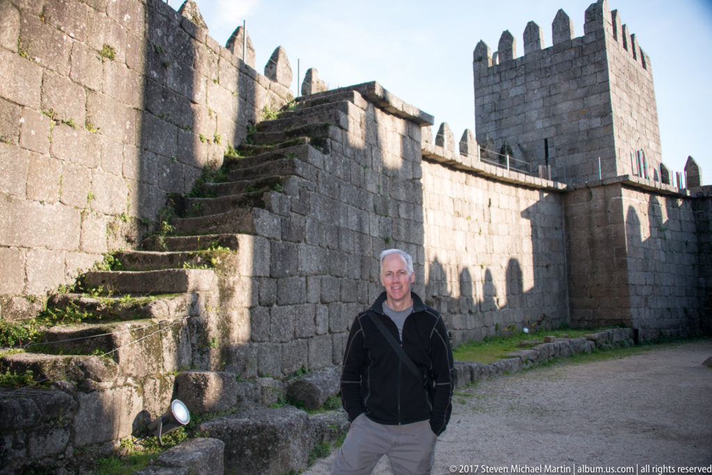 Man standing beside Guimarães Castle wall and steep exterior stone stair, with crenellations, arched openings, and a square tower behind.