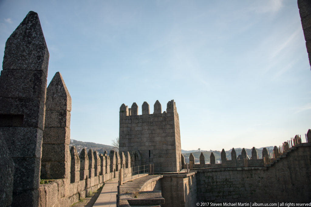Granite wall walk with pointed crenellations leading toward a square battlemented tower at Guimarães Castle.