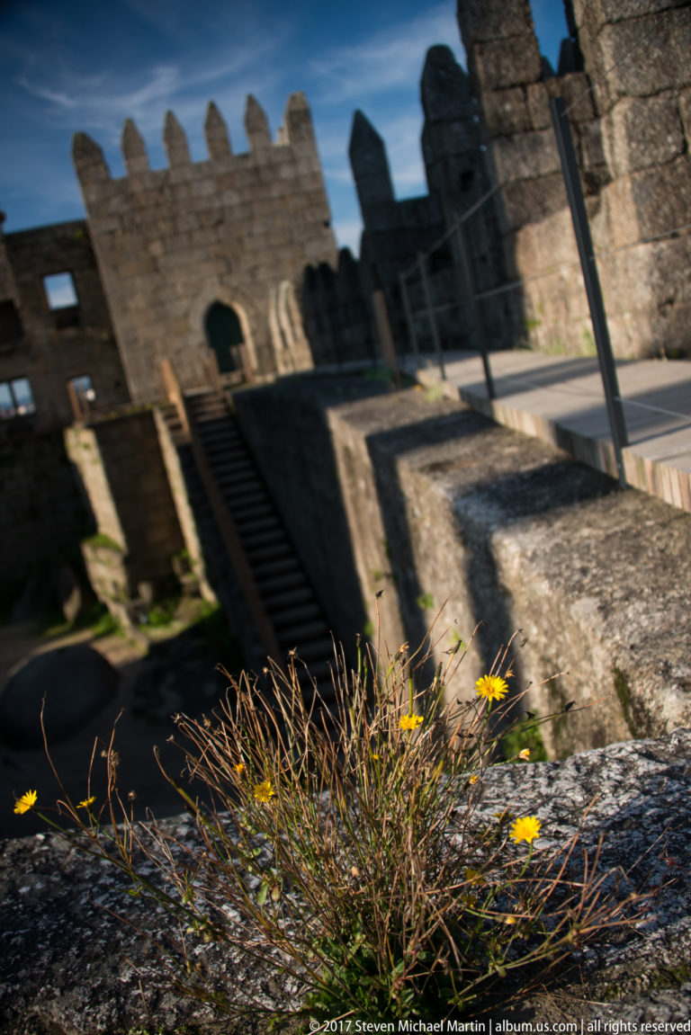 Angled view across Guimarães Castle interior with steep stairs, crenellated walls, arched openings, and yellow flowers in the foreground.