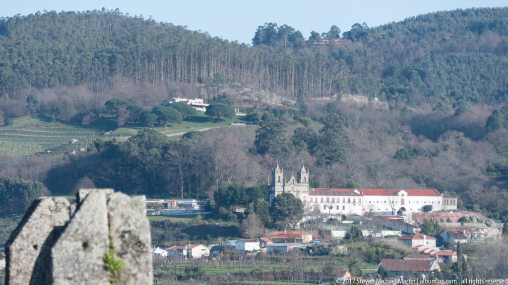 Distant white monastery and church complex on a wooded hillside, viewed past a pointed stone battlement from Guimarães Castle.
