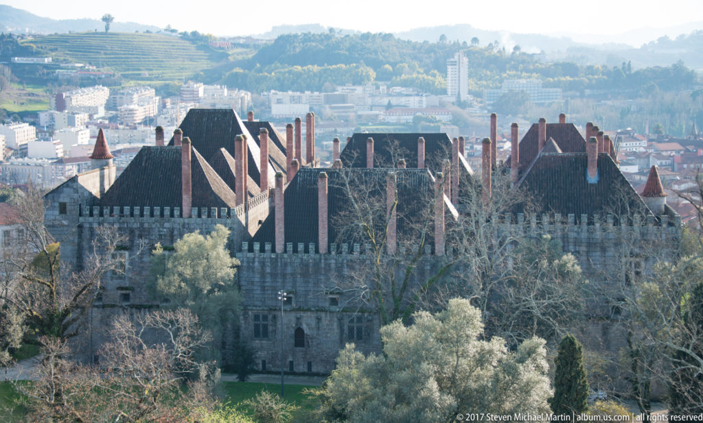 Palace of the Dukes of Braganza seen from above, with crenellated walls, steep roofs, tall brick chimneys, and Guimarães beyond.
