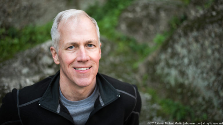 Close portrait of a smiling gray-haired man in a black jacket with blurred stone walls and greenery behind him.