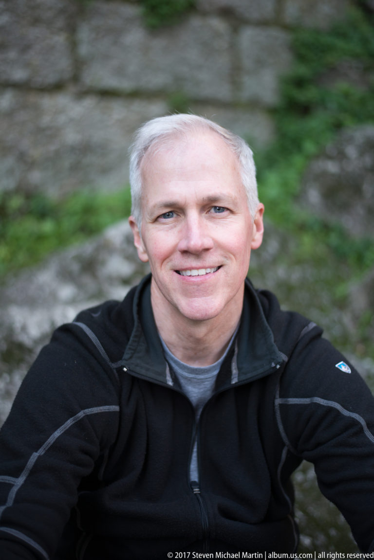 Smiling gray-haired man in a dark jacket photographed at close range against blurred stone and greenery.