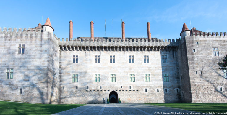Broad granite façade of the Palace of the Dukes of Braganza with corner towers, crenellations, tall chimneys, and central arched entrance.