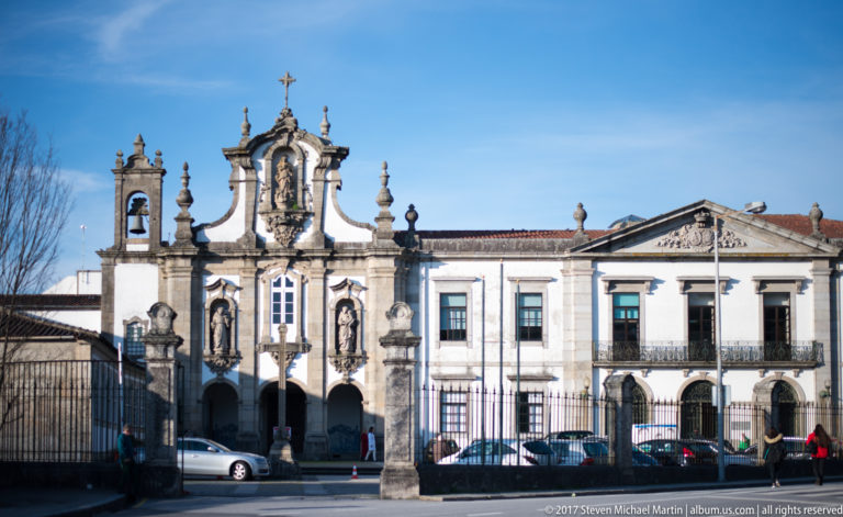 Ornate chapel façade with statues, bell opening, pinnacles, and cross attached to a long white building with balconies and arcades.