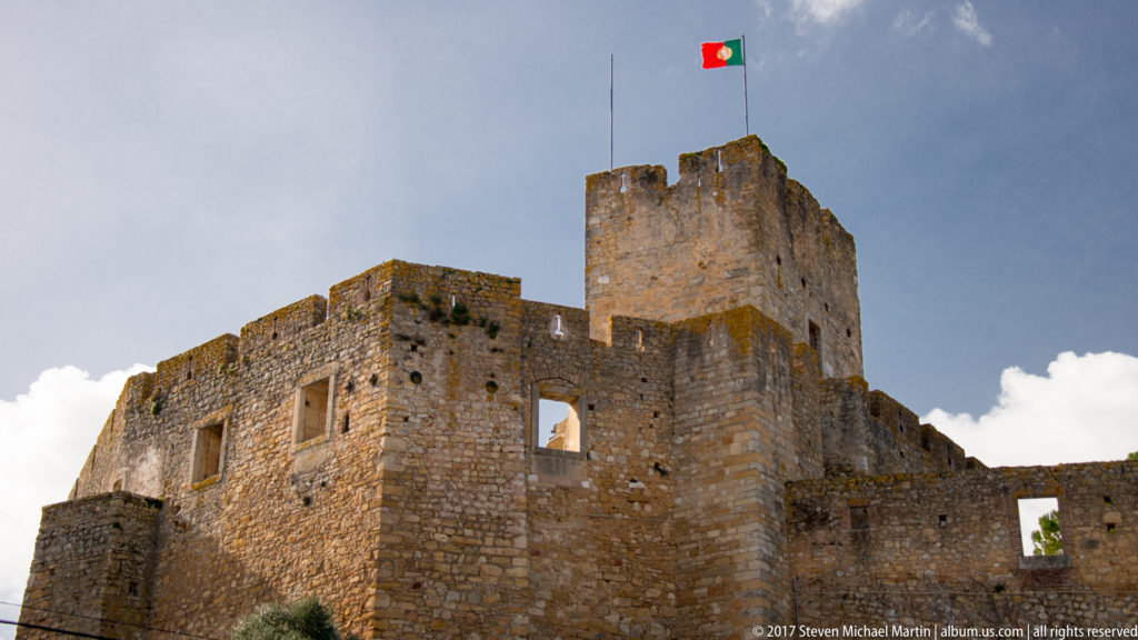 Tall rectangular castle tower flying the Portuguese flag above lower stone walls and openings beneath a dark blue-gray sky.