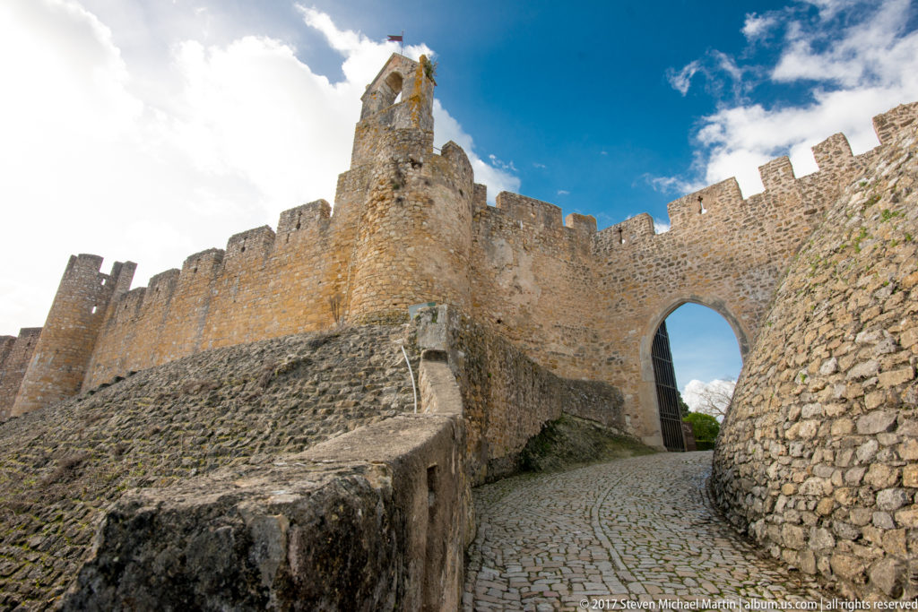Cobbled path curving uphill through a tall arched gateway in crenellated stone castle walls with a small tower above.