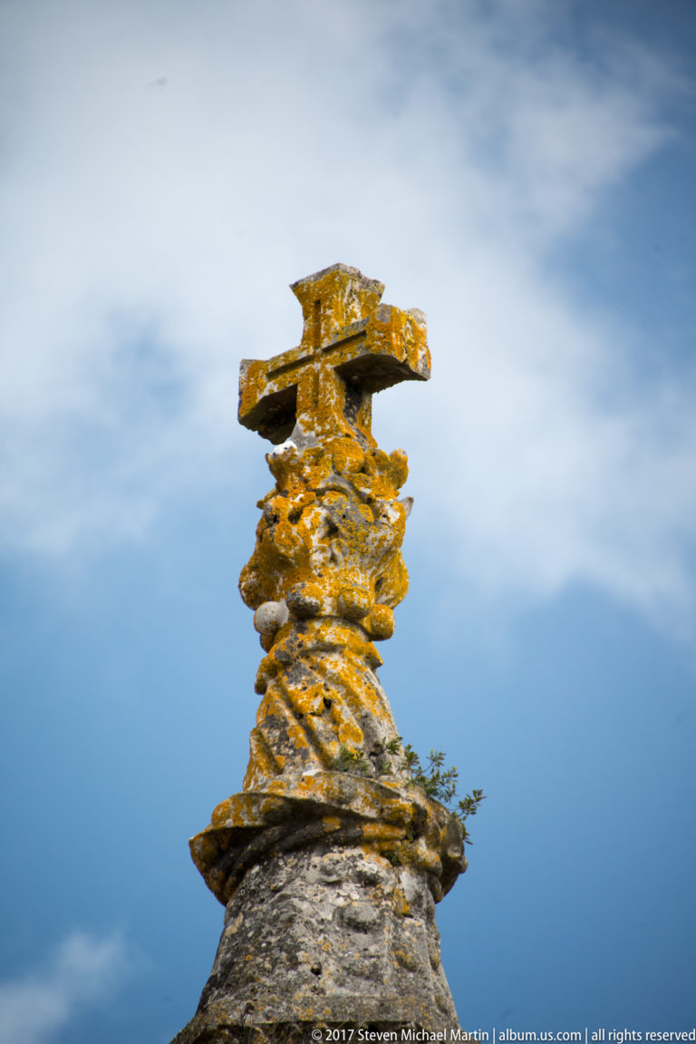Weathered carved stone pinnacle topped by a cross, with golden lichen against a cloudy blue sky.