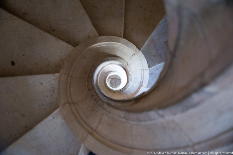Pale stone spiral staircase curling upward around a smooth central rail toward a bright opening.