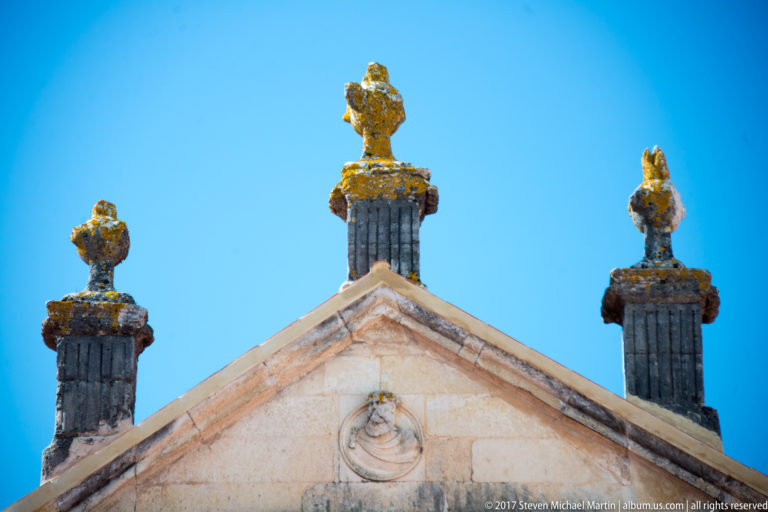 Three weathered sculpted stone finials rise above a pale triangular gable against a blue sky.