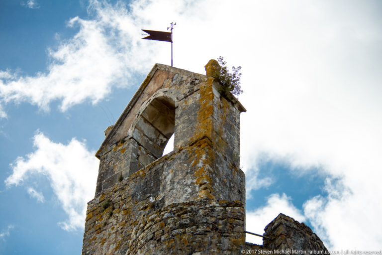 Weathered stone bell gable with an empty arched opening, lichen, small plants, and a metal weather vane against clouds.