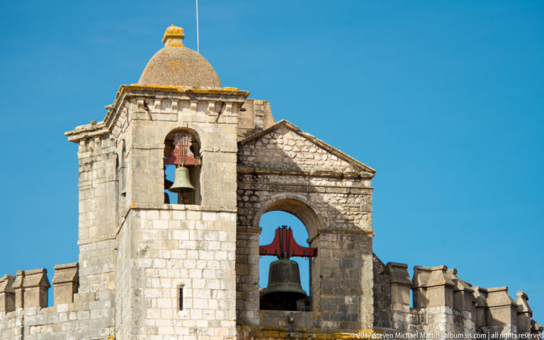Two adjoining stone bell openings with bronze bells and red supports, one beneath a domed tower and one in a gabled arch.