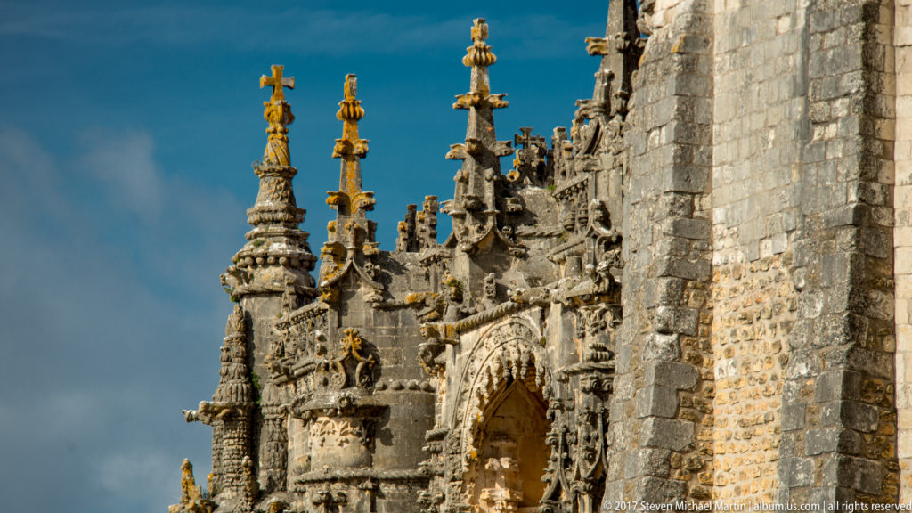 Oblique low-angle view of an ornate pinnacled church portal beside a tall plain stone wall under a blue sky.
