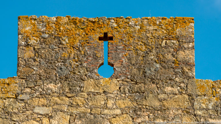 Close view of a stone battlement pierced by a narrow vertical slit ending in a round opening, with lichen along the top.