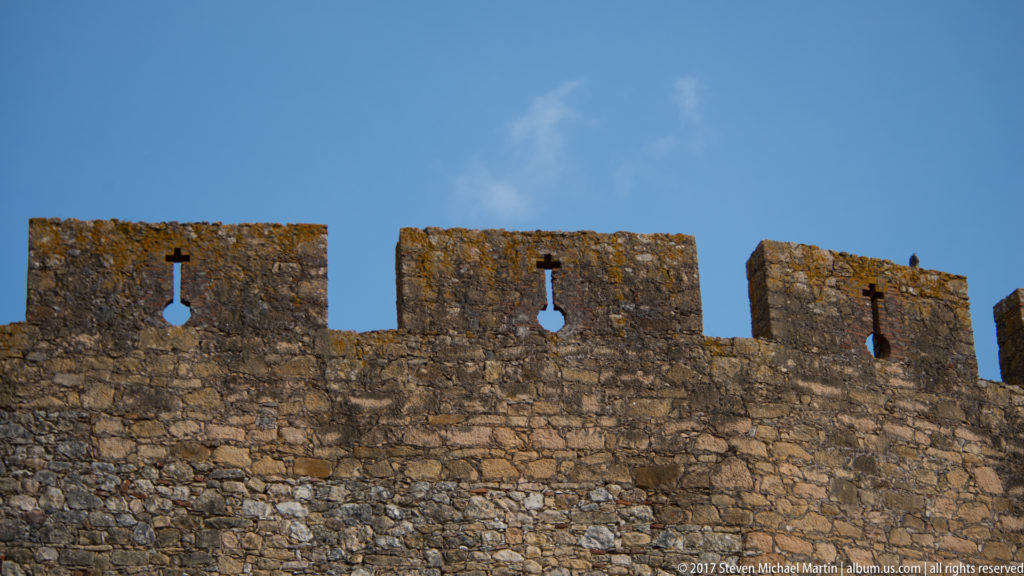 Low view of stone castle battlements with repeated merlons pierced by narrow vertical slots ending in circular openings.