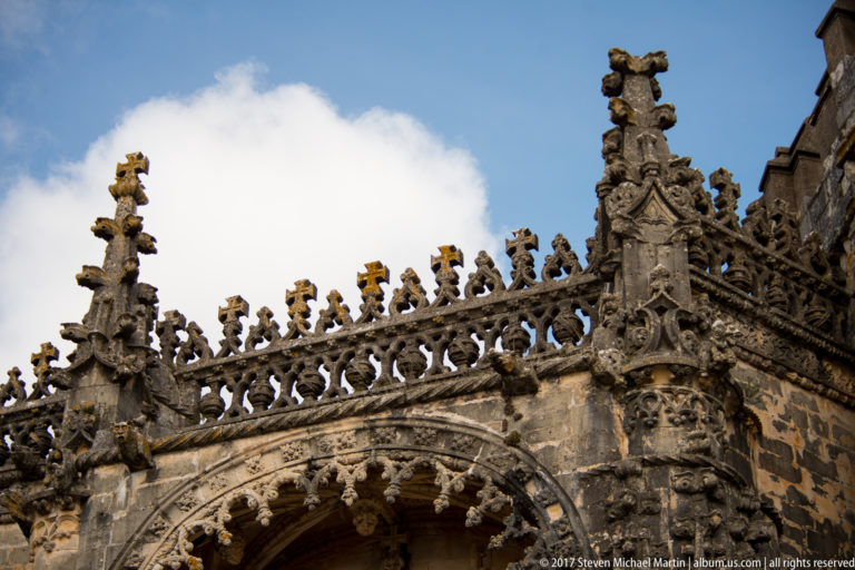 Close upward view of a carved stone parapet with pinnacles, pierced tracery, crosses, and an ornate arched opening below.